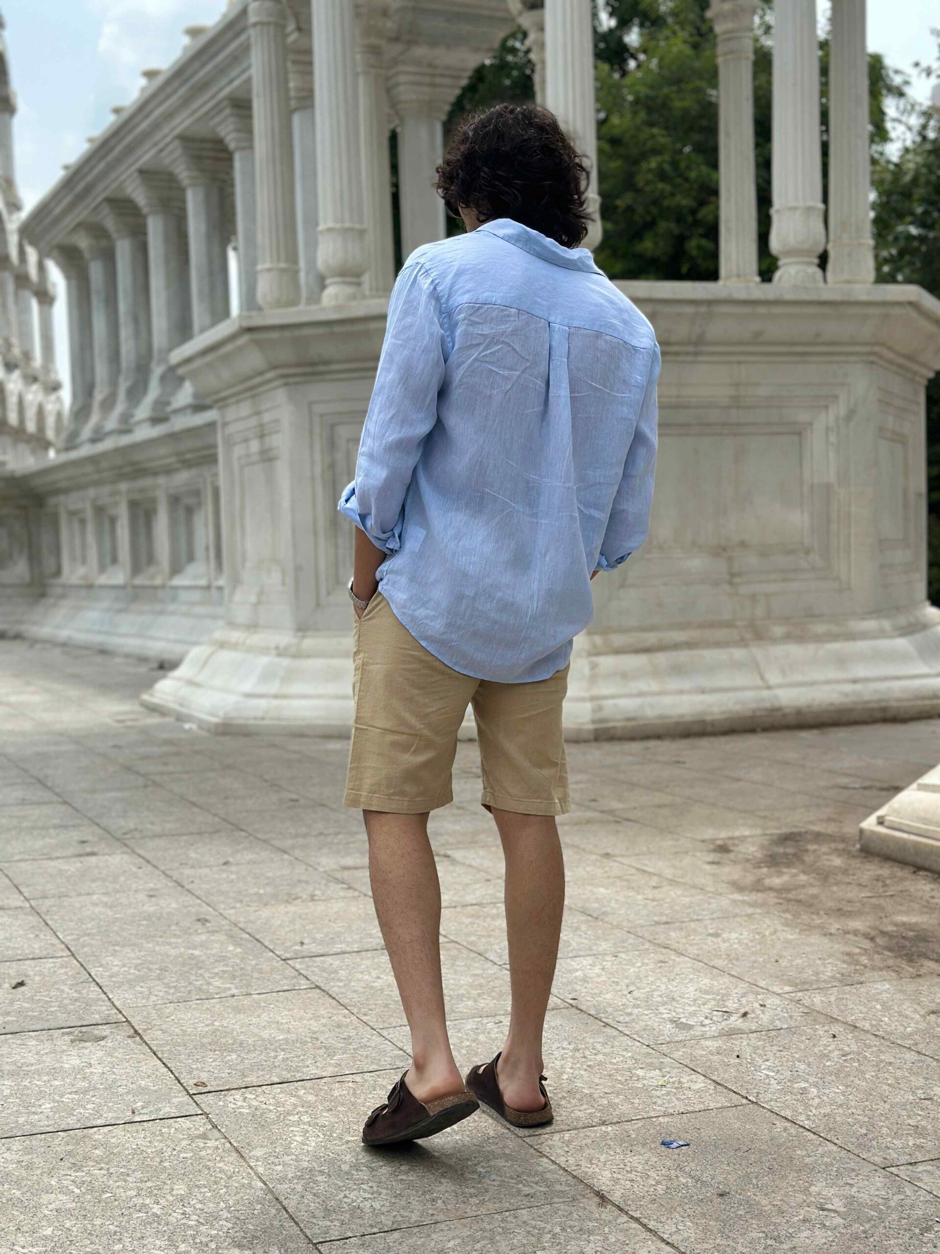 Back view of a man in cream linen shorts and a light blue shirt, walking away.